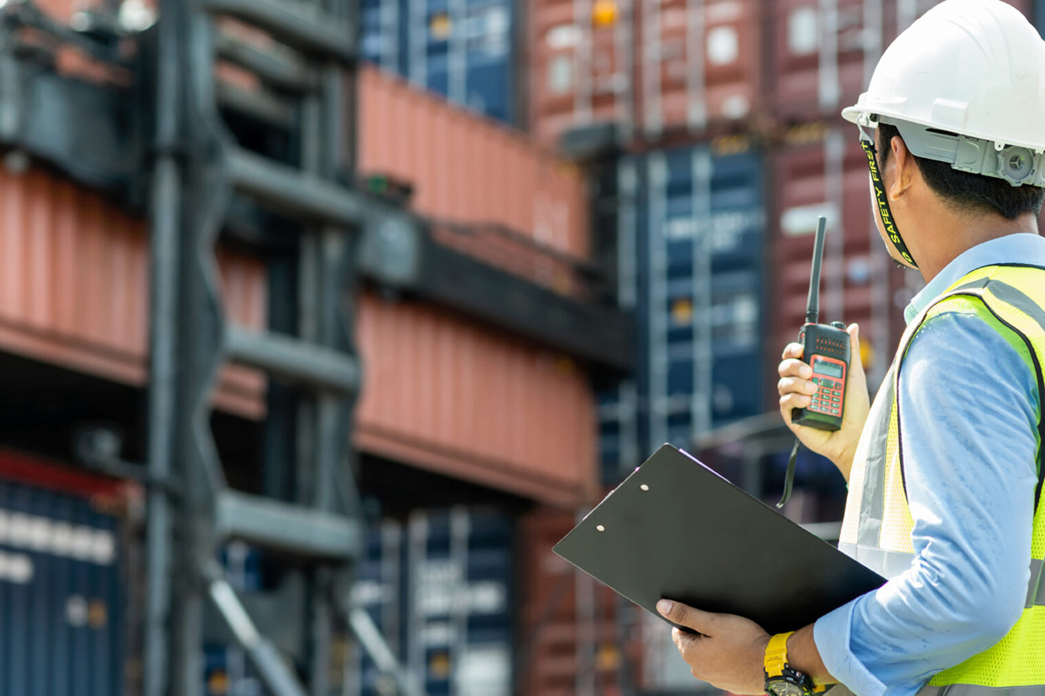 Ein Arbeiter steht mit einem Walkie-Talkie vor vielen Containern. Ein Mann mit Warnweste und Bauarbeiterhelm steht vor Containern mit einem Klemmbrett und einem Walkie-Talkie in der Hand.