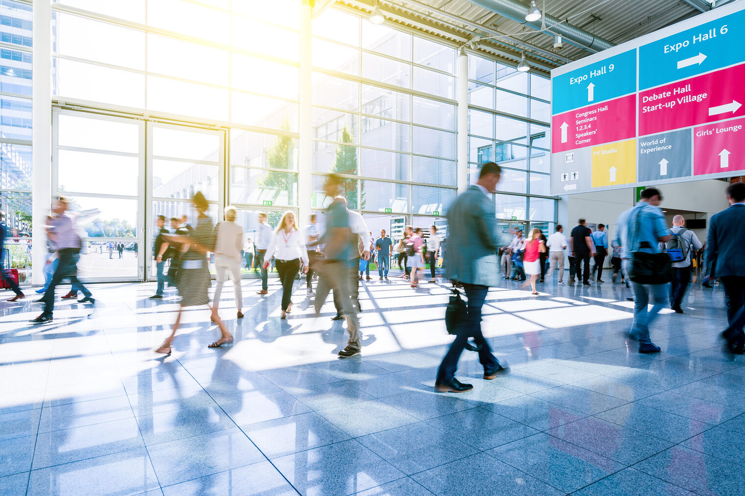 Menschenmenge in einem hellen Messe- oder Kongresszentrum, im Hintergrund große Fensterfront und farbige Wegweiser.