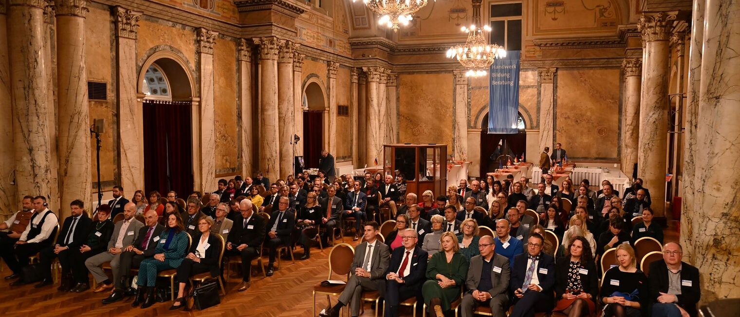 Menschen sitzen im gro&szlig;en, festlichen Saal und h&ouml;ren zu. 