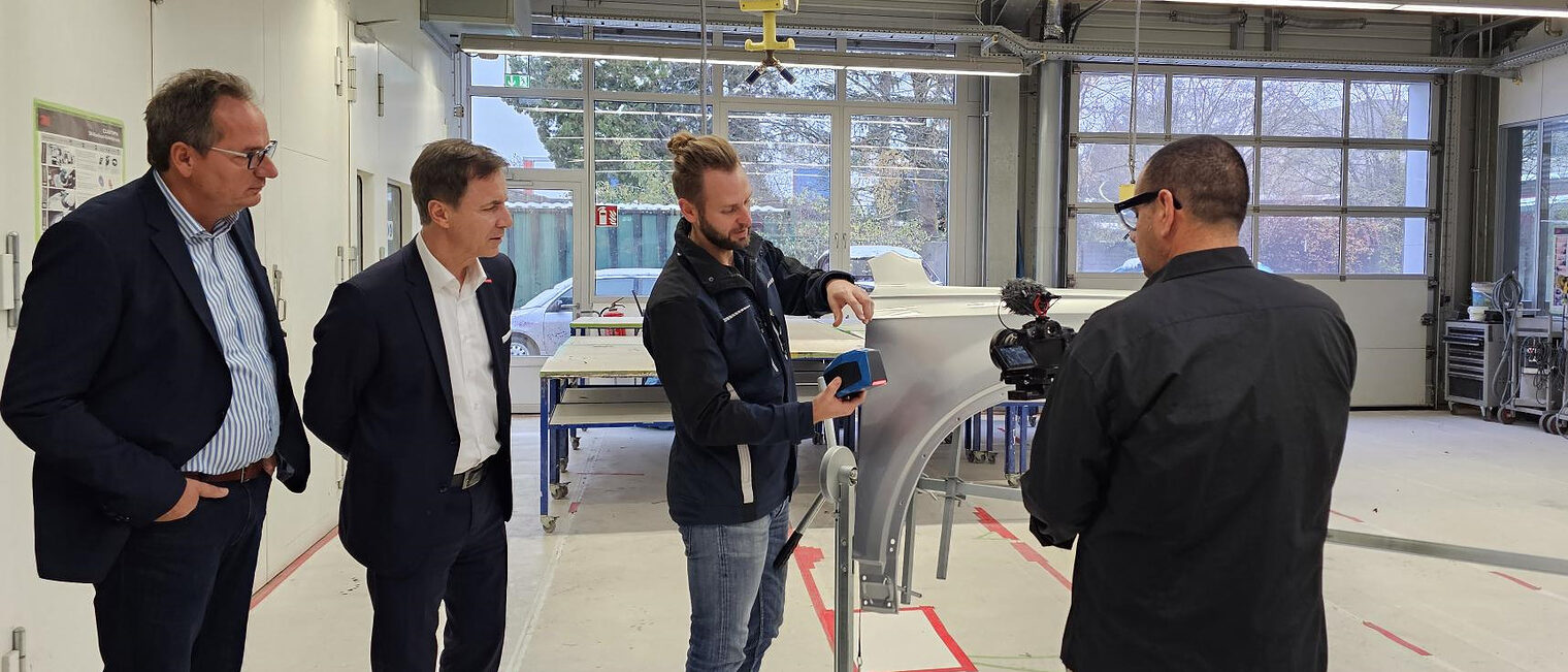 PPG-Trainingsmanager Dirk Czub (links) und stv. HWK-Hauptgeschäftsführer Hans Schmidt (2.v.li.) bei der Demonstration der automatischen Farbtonausmessung. Männer schauen sich interessiert die neue Farbmischanlage an. Ein Filmer nimmt Aufzeichnung von der Maschine auf.