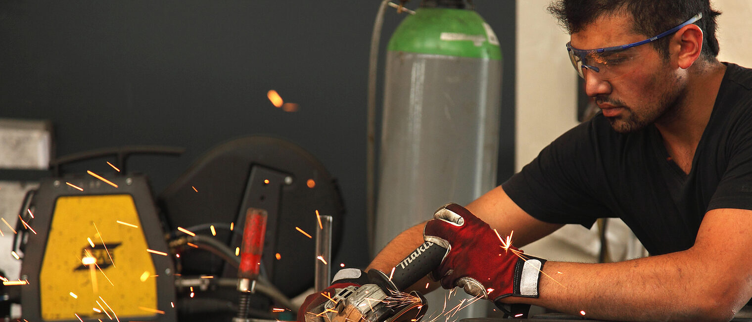 Ein Mann mit Schutzbrille und Handschuhen arbeitet gewissenhaft mit einer Flex in einer Werkstatt, dabei sprühen Funken. 