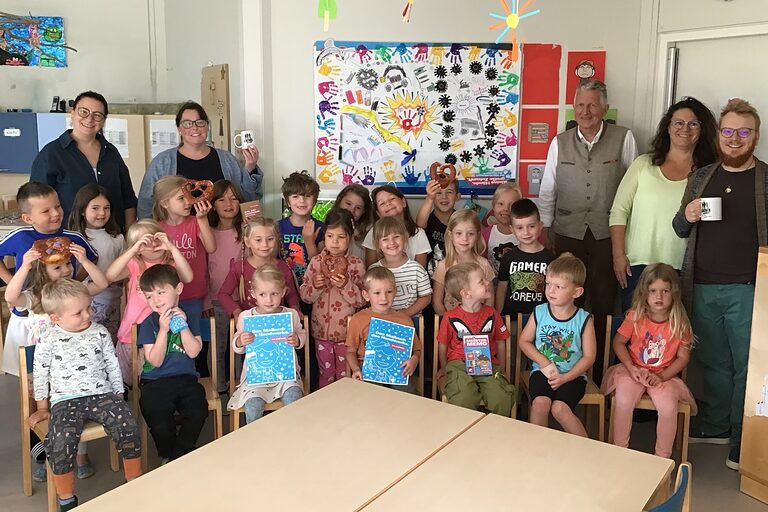 Kindergartenkinder, Erwachsene und HWK-Vizepräsident Hans Kastl sind in einem Raum mit bunter Wanddekoration. Einige Kinder halten Brezeln und Hefte.