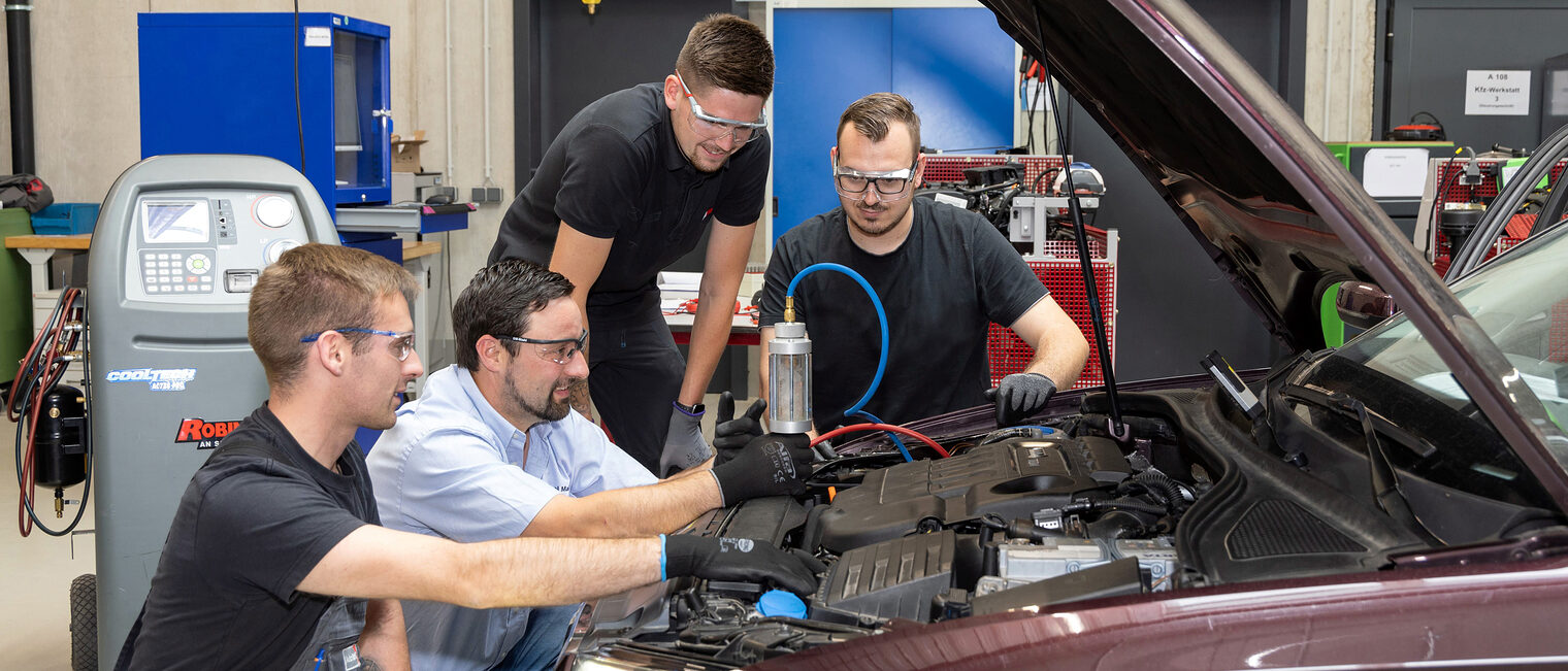 Drei Auszubildende im Kfz-Mechatronikerhandwerk untersuchen mit ihrem Ausbilder ein Auto in der KFZ-Werkstatt.