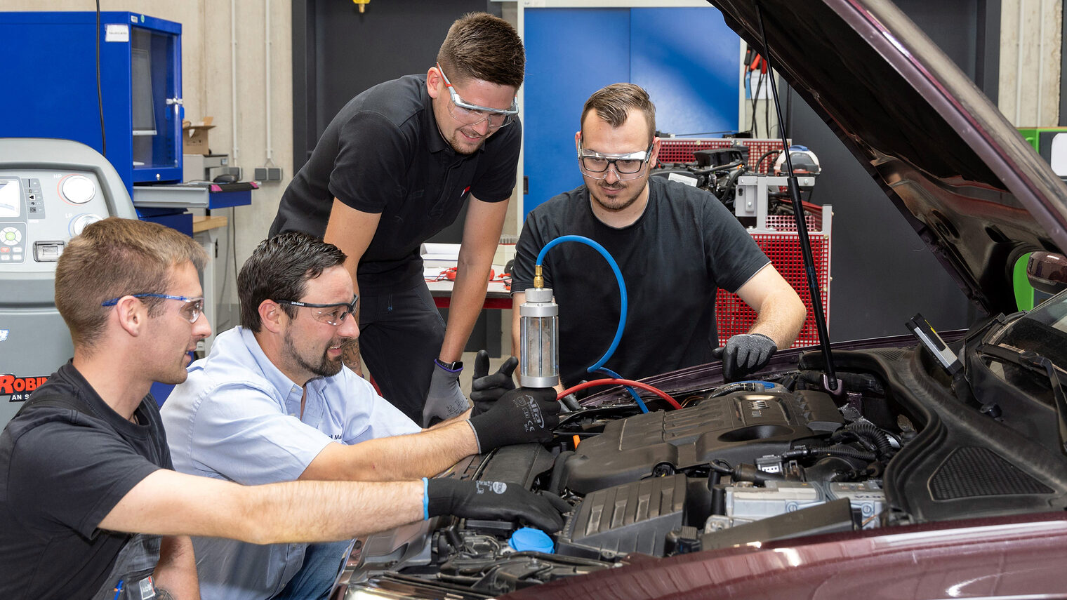 Drei Auszubildende im Kfz-Mechatronikerhandwerk untersuchen mit ihrem Ausbilder ein Auto in der KFZ-Werkstatt.