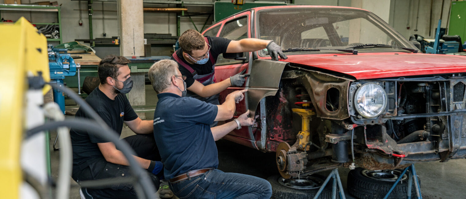 Zwei Meisterschüler befestigen mit dem Ausbilder ein Teil an einer Auto-Karrosserie.