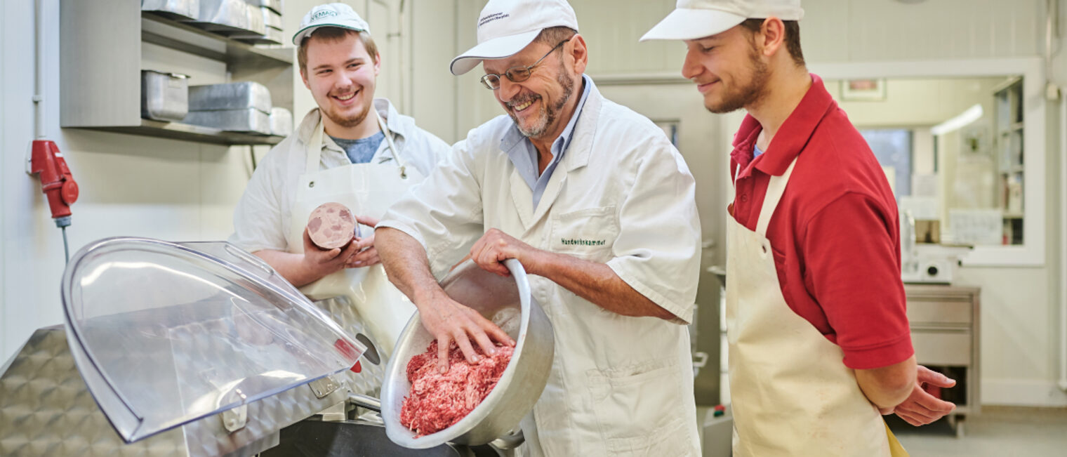 Ein Ausbilder füllt Hackfleisch in einen Mischer, zwei Auszubildende sehen ihm dabei zu.