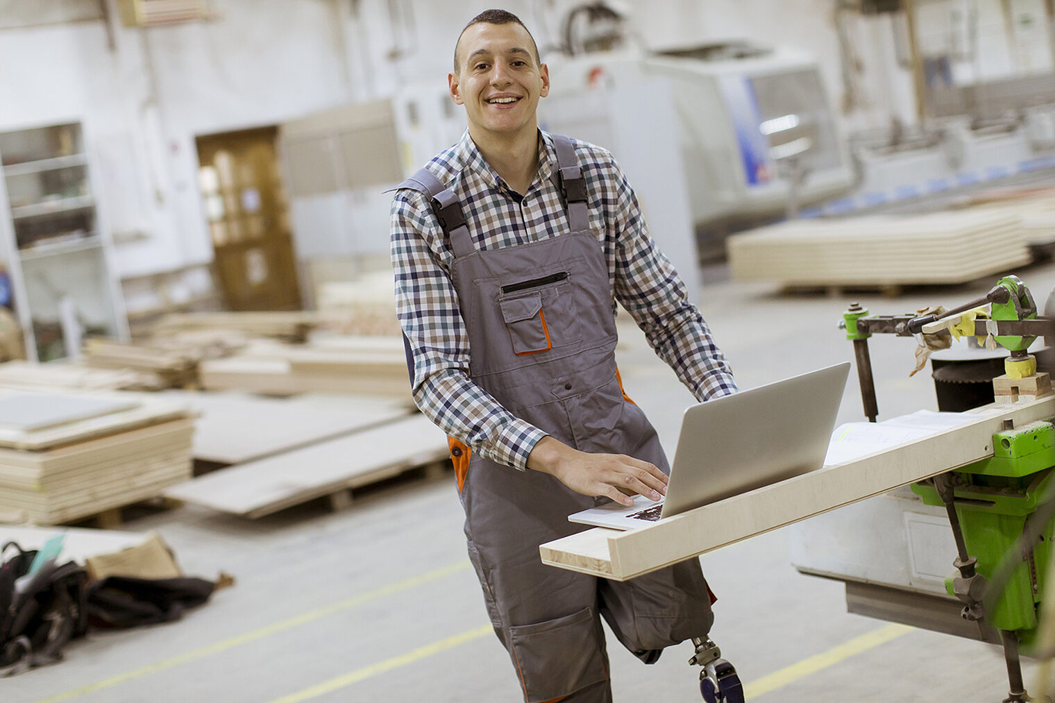 Ein Mann mit einer Beinprothese steht mit dem aufgeklappten Laptop in der Werkstatt. 