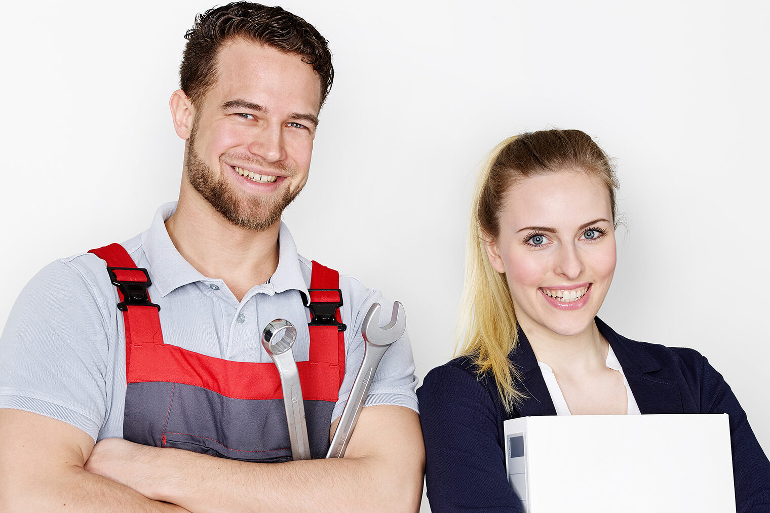 Ein junger Handwerker und eine junge Frau stehen selbstbewusst vor der Kamera. Er mit einem Schraubenschlüssel und sie mit einem Ordner. 