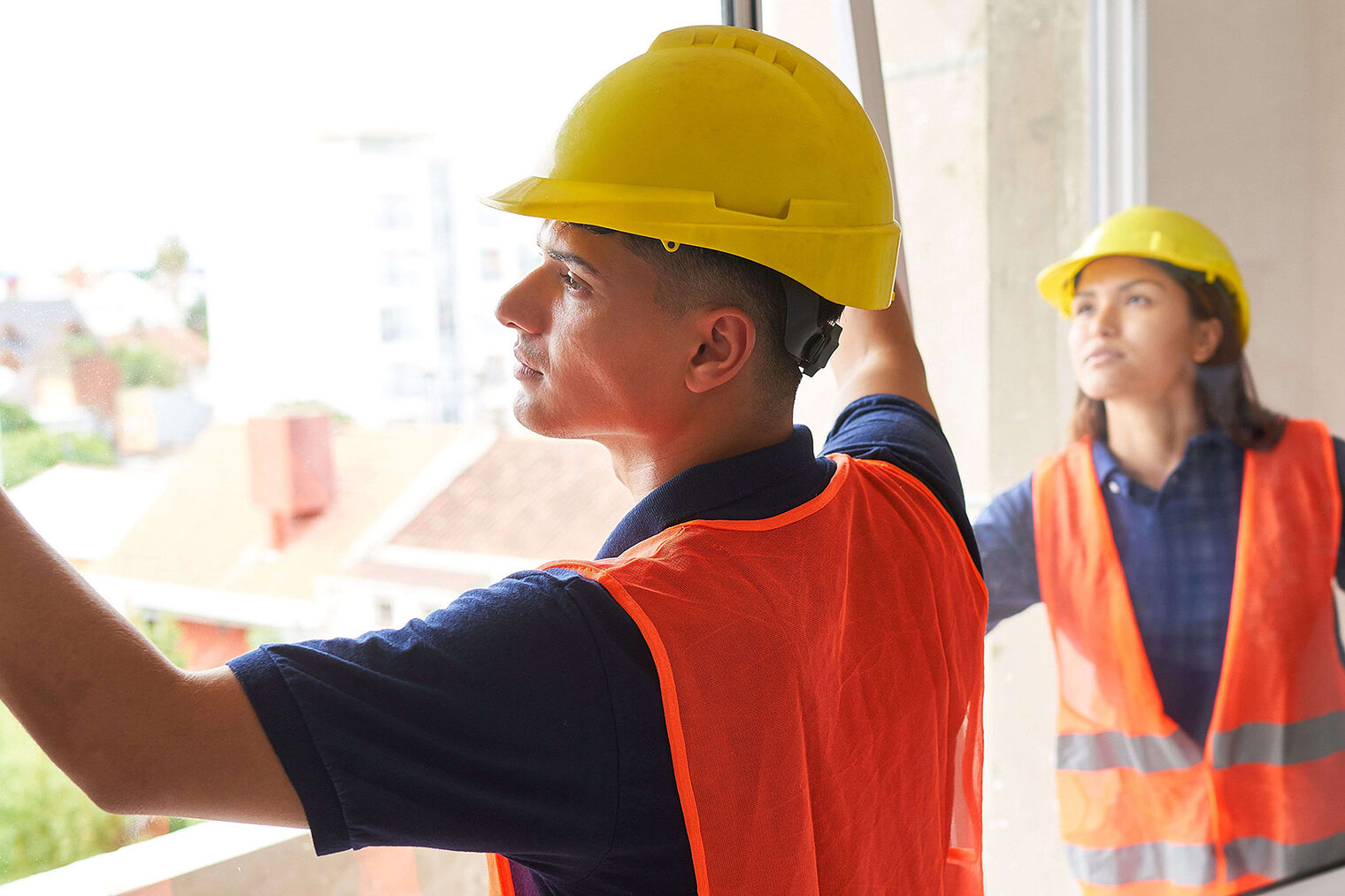 Ein Fensterbauer und eine Fensterbauerin montieren eine Balkontür in einem Neubau. Sie tragen Warnwesten und Helme.