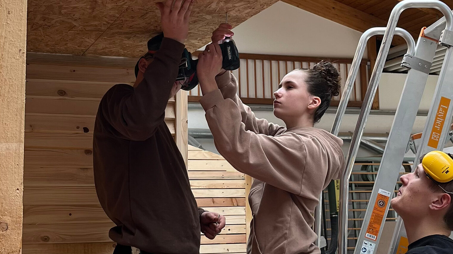 Ein Mädchen mit Bohrmaschine steht auf einer Leiter und arbeitet an einer Holzdecke; zwei Männer daneben geben Hilfestellung.